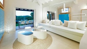 A modern living room with a white sofa, a coffee table, and a dining area with chairs and a table. There is a view of greenery outside. - Lovely apartment in villa for 6 guests with A/C, pool, TV and terrace (San Teodoro)