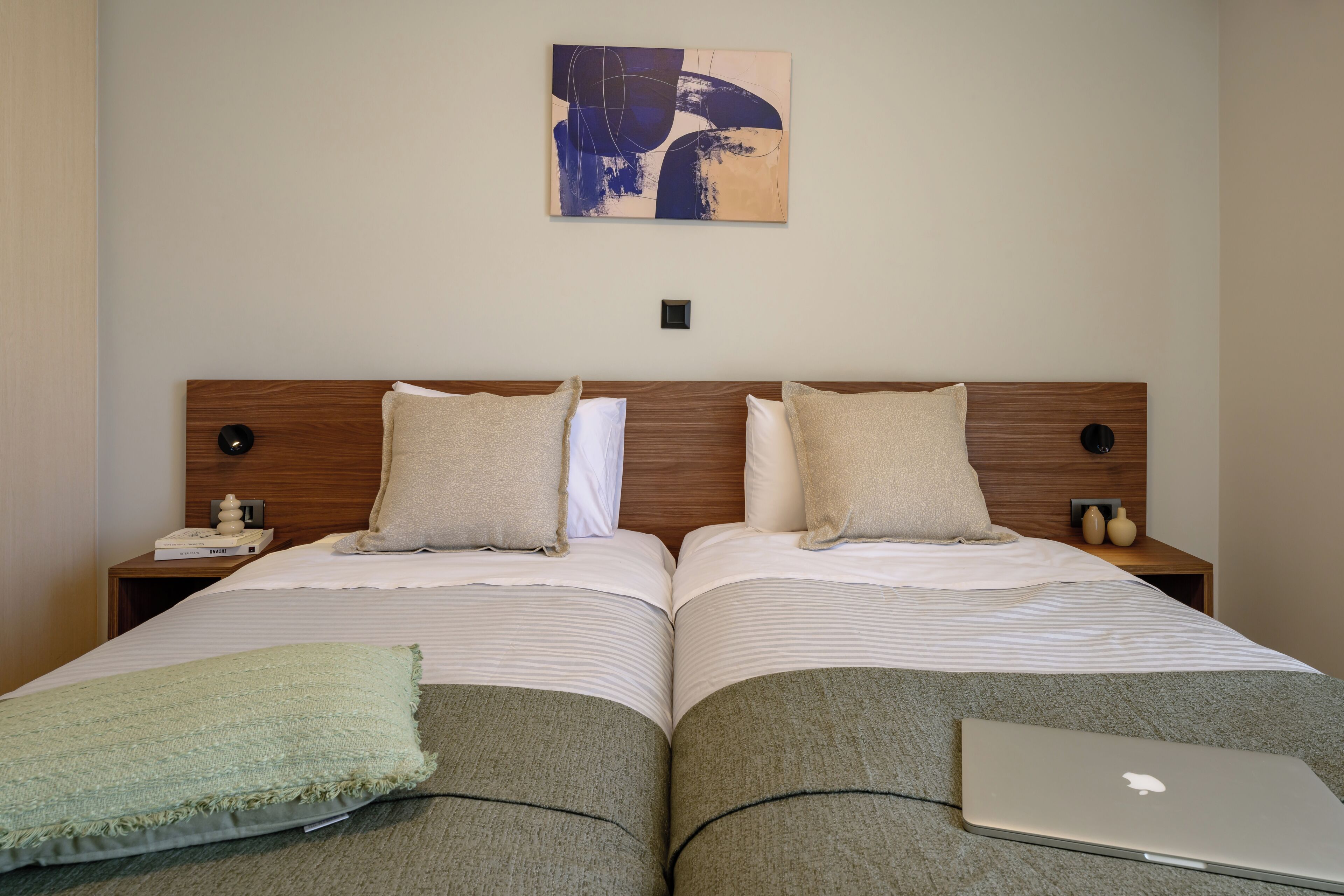 A double bed with two pillows, a bedside table with a lamp, a wall art piece, and a laptop on the bed.