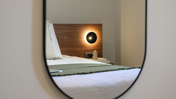 A bedroom with a wooden headboard, a bedside table with a lamp, and a mirror reflecting the room. - Elise Apartment Airport  by Airstay (Spata-Artemida)