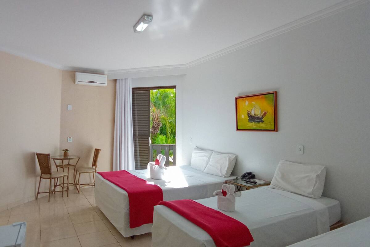 A hotel room with two beds, a TV, a desk with chairs, and a window with a view of greenery.