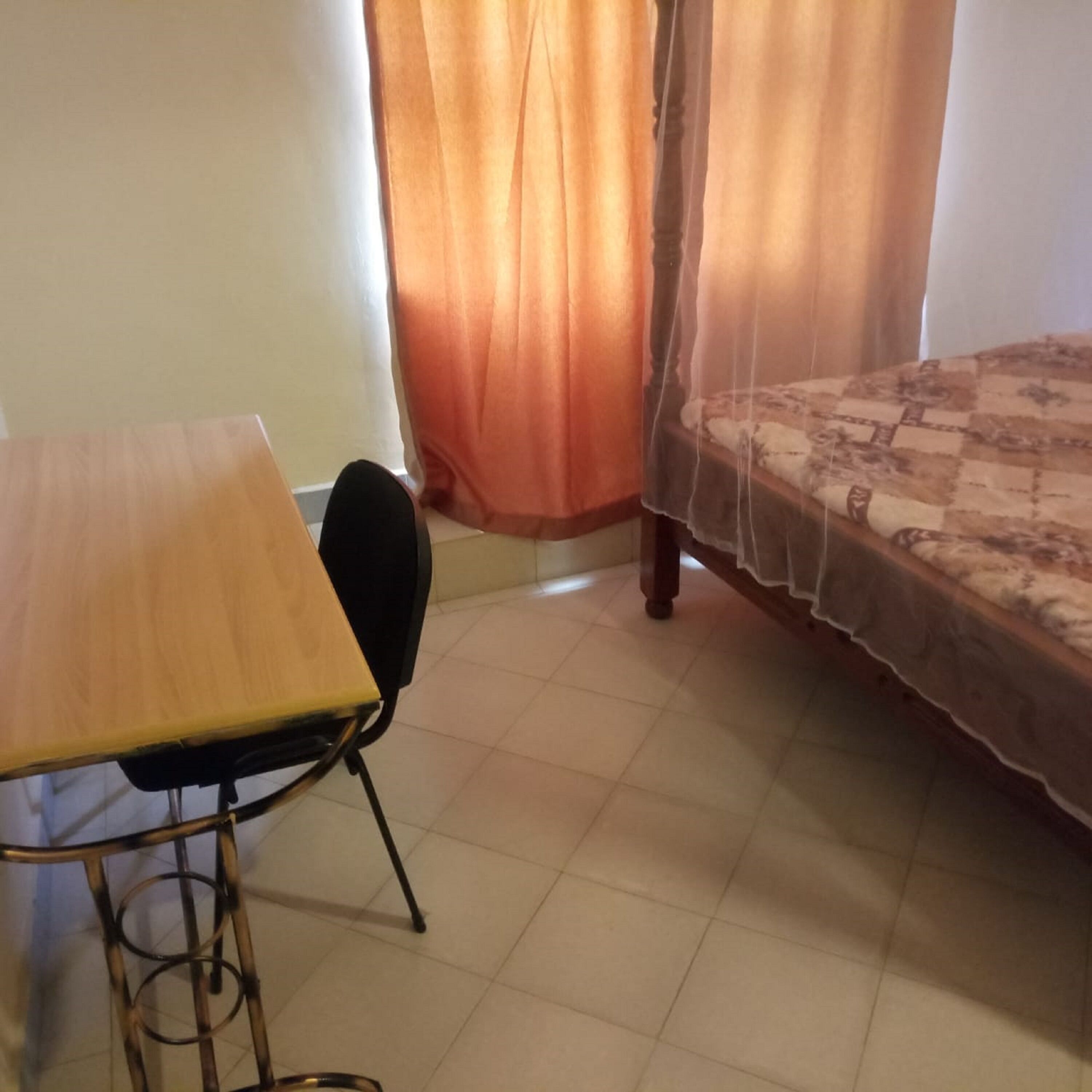 Une pièce avec un lit recouvert d’une couvre-lit à motifs, une table en bois avec une chaise, et une fenêtre ornée de rideaux orange.