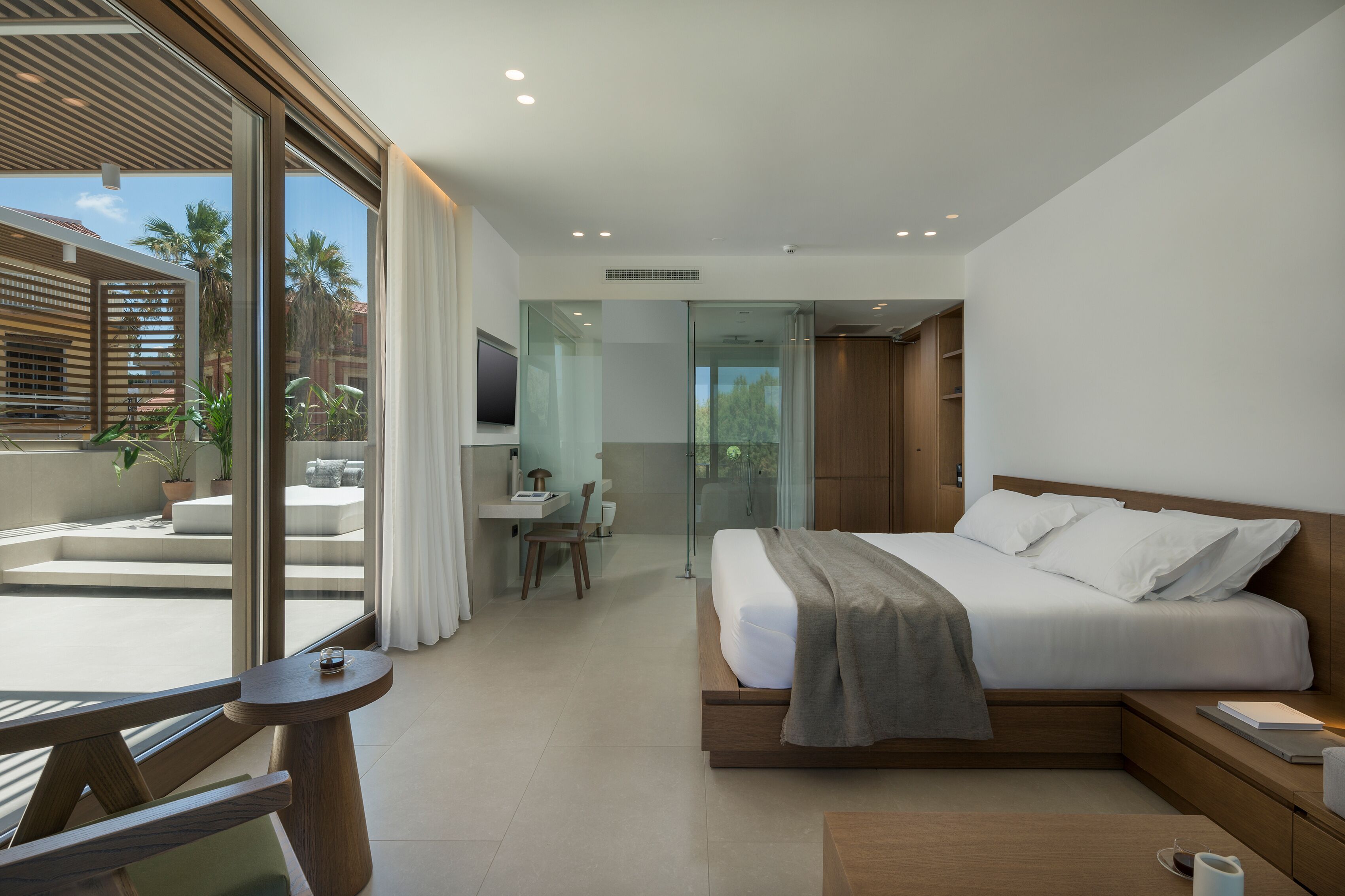 A modern hotel room with a large bed, a desk, and a balcony with plants.