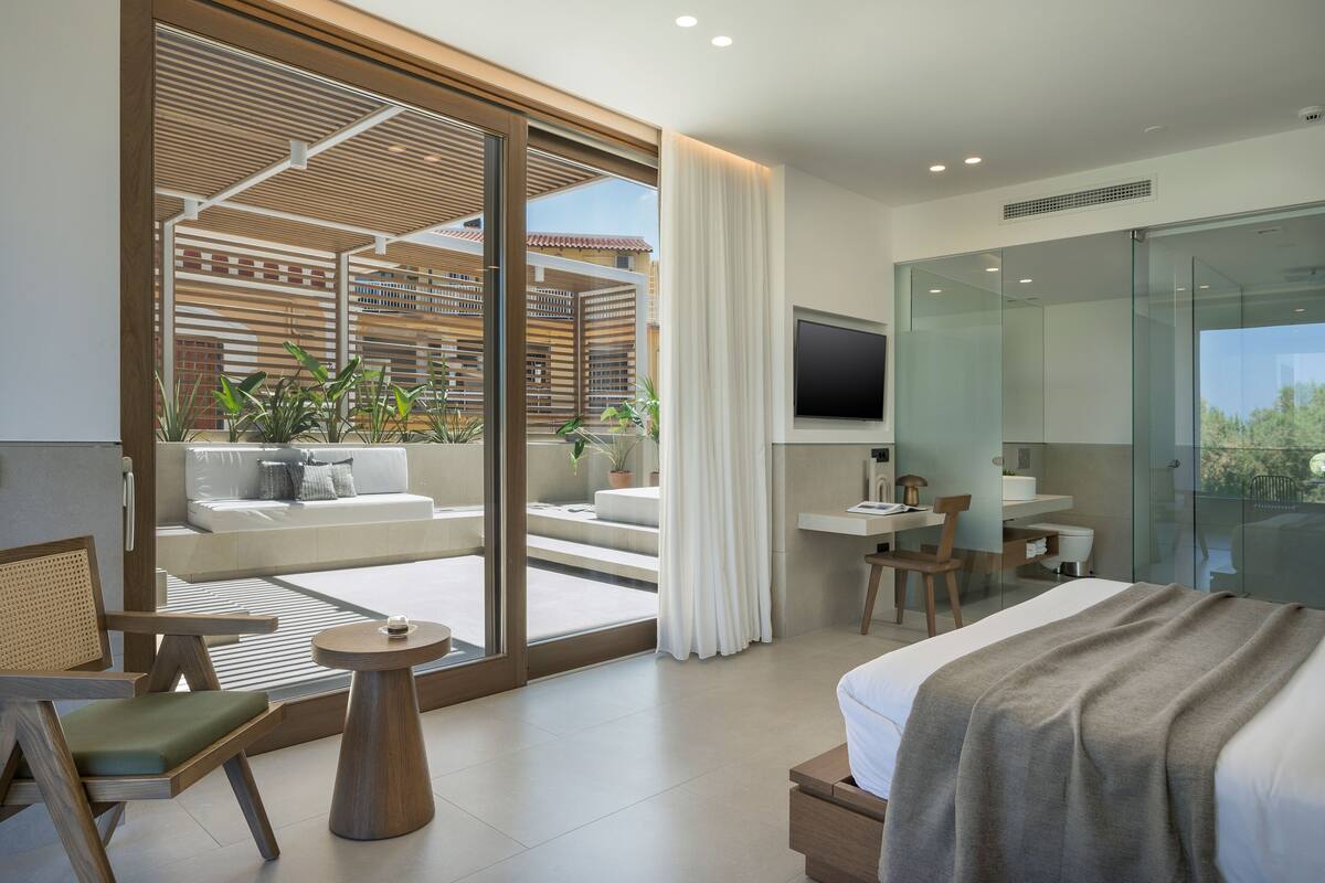 Ein modernes Hotelzimmer mit einer großen Glasschiebetür, die auf einen Balkon führt, einem Bett mit grauer Decke, einem Schreibtisch mit Stuhl und einem an der Wand montierten Fernseher.