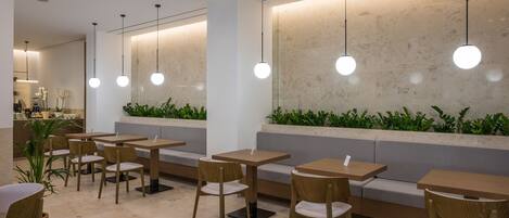 A modern cafe with wooden tables and chairs, a counter area, and green plants along the walls.