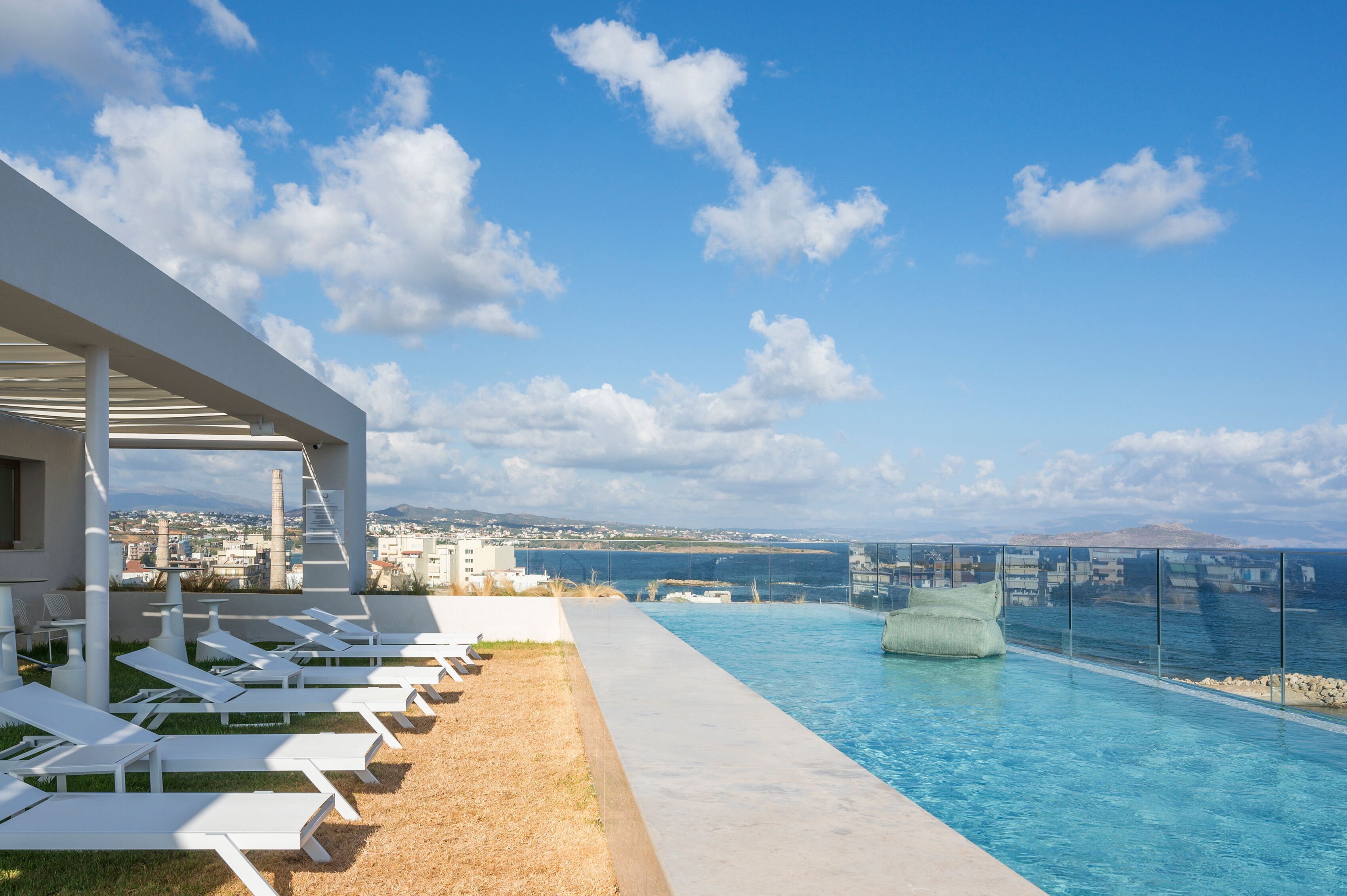 Uma área de piscina moderna com espreguiçadeiras e vista desobstruída para o mar e a paisagem urbana ao longe.