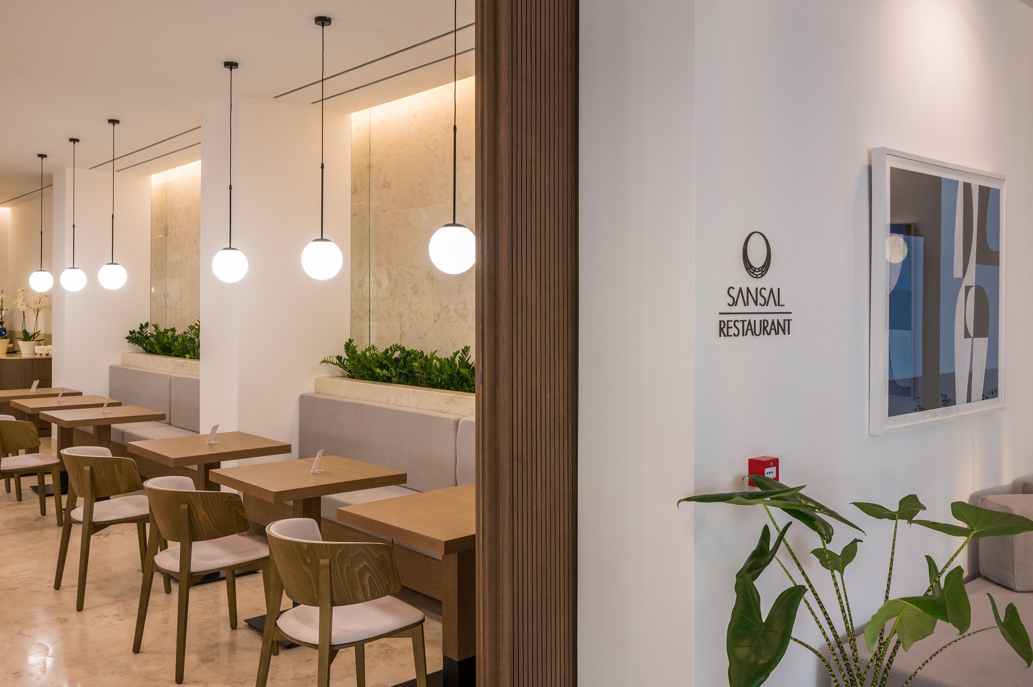 L’intérieur d’un restaurant, avec des tables et des chaises en bois, des lampes suspendues et un panneau fixé au mur sur lequel on peut lire : « RESTAURANT SANSAL ».