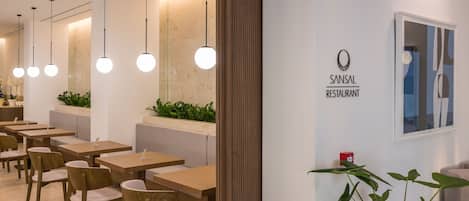 L’intérieur d’un restaurant, avec des tables et des chaises en bois, des lampes suspendues et un panneau fixé au mur sur lequel on peut lire : « RESTAURANT SANSAL ».