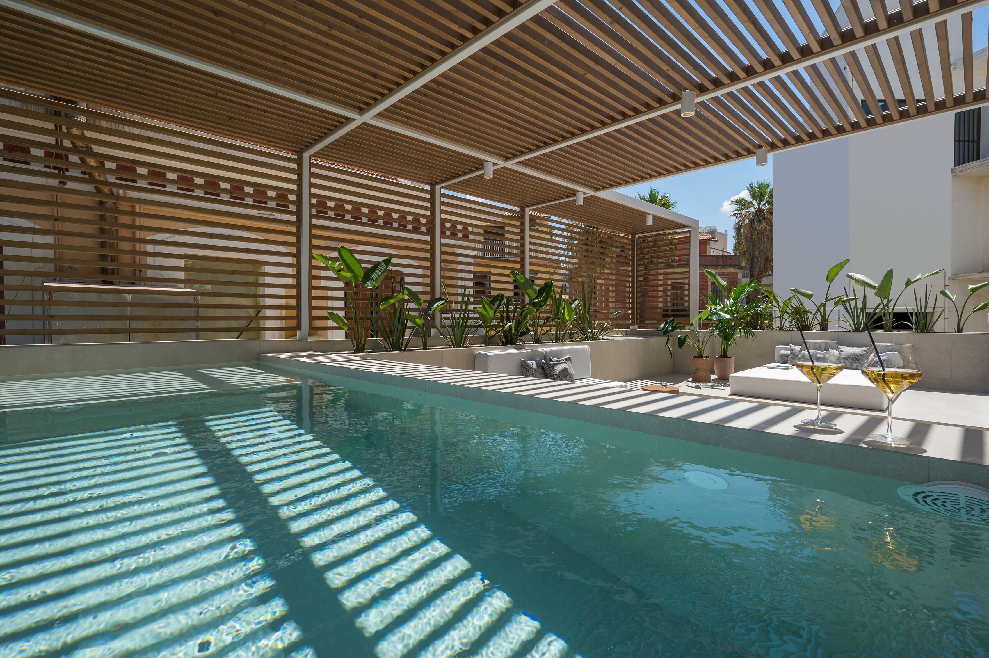 A swimming pool with a wooden pergola overhead, surrounded by potted plants and outdoor seating.