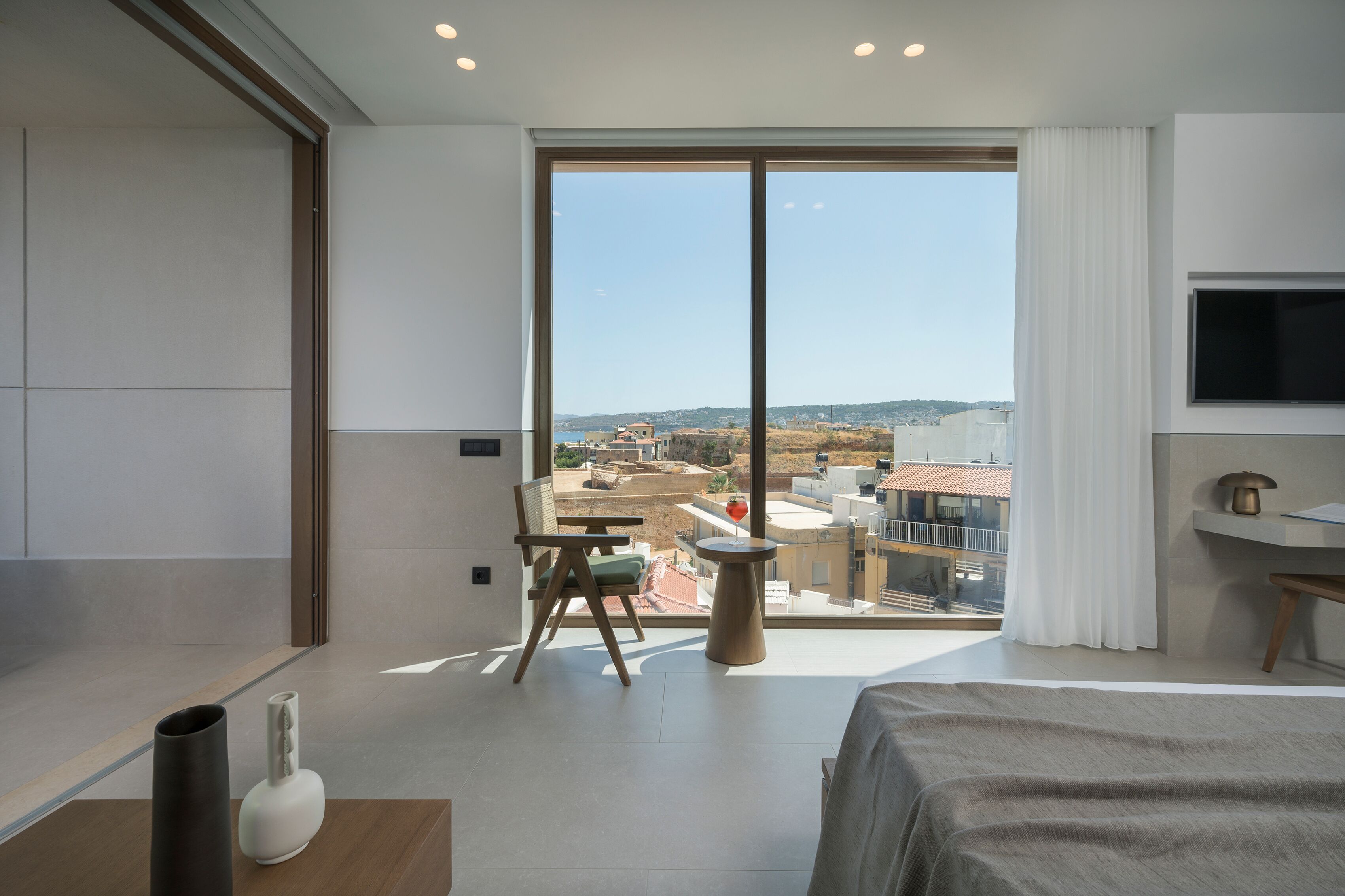 A modern hotel room with a large window, a bed, a TV, and a small table with a chair.