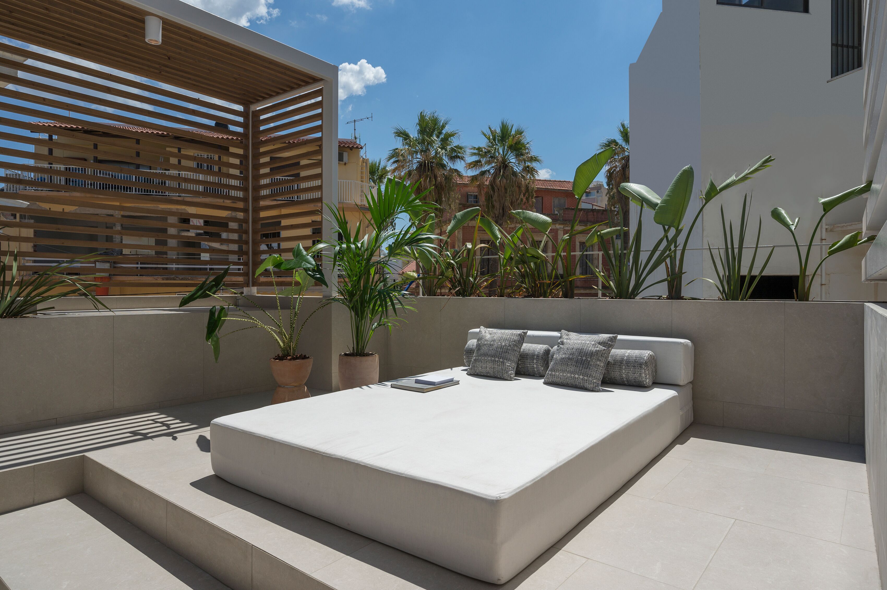 Un espacio exterior moderno con zona de estar, un sofá blanco y cojines, rodeado de plantas en macetas y una estructura de listones de madera.