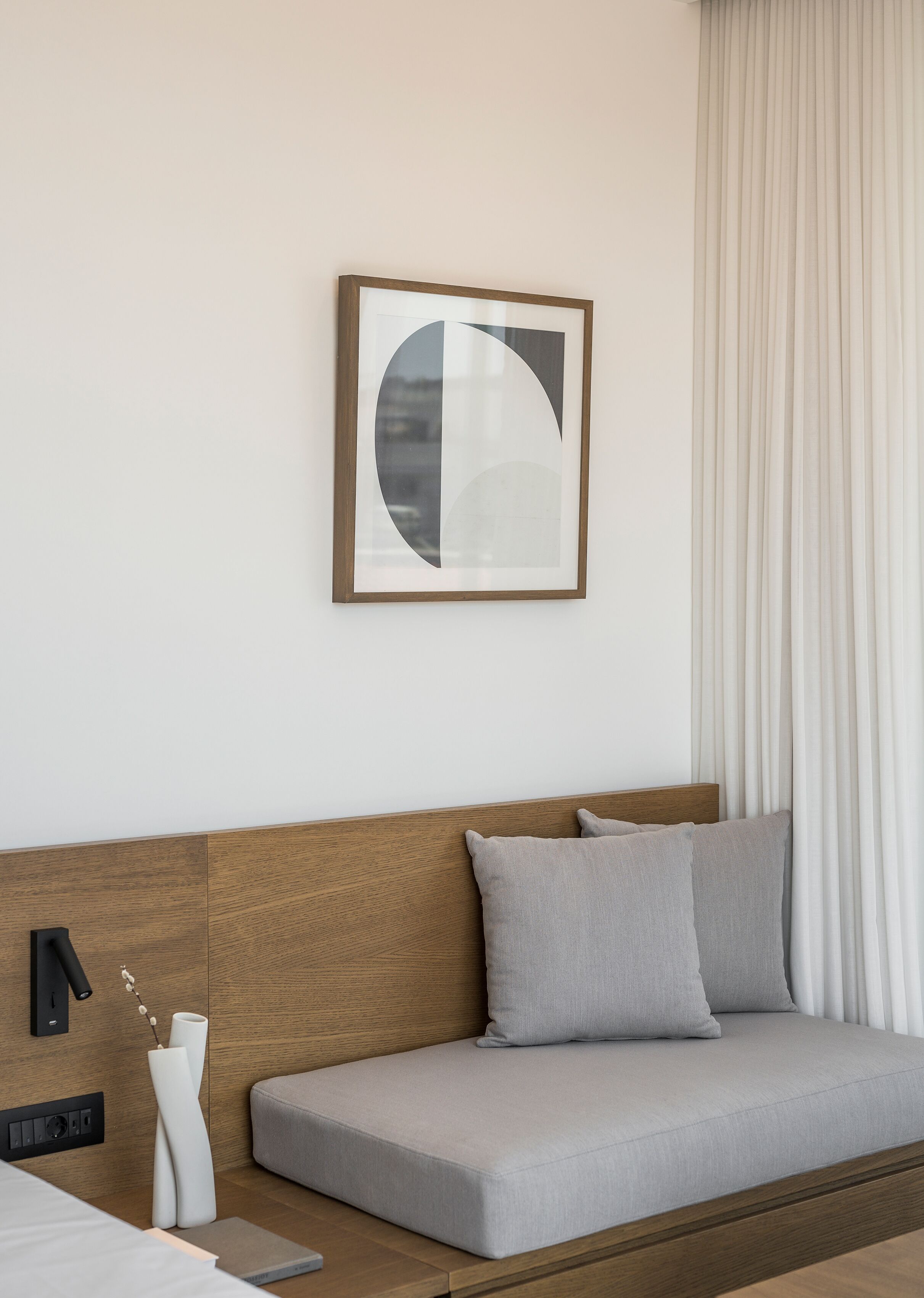 A modern hotel room with a wooden headboard sofa, a framed abstract artwork, and a white curtain.