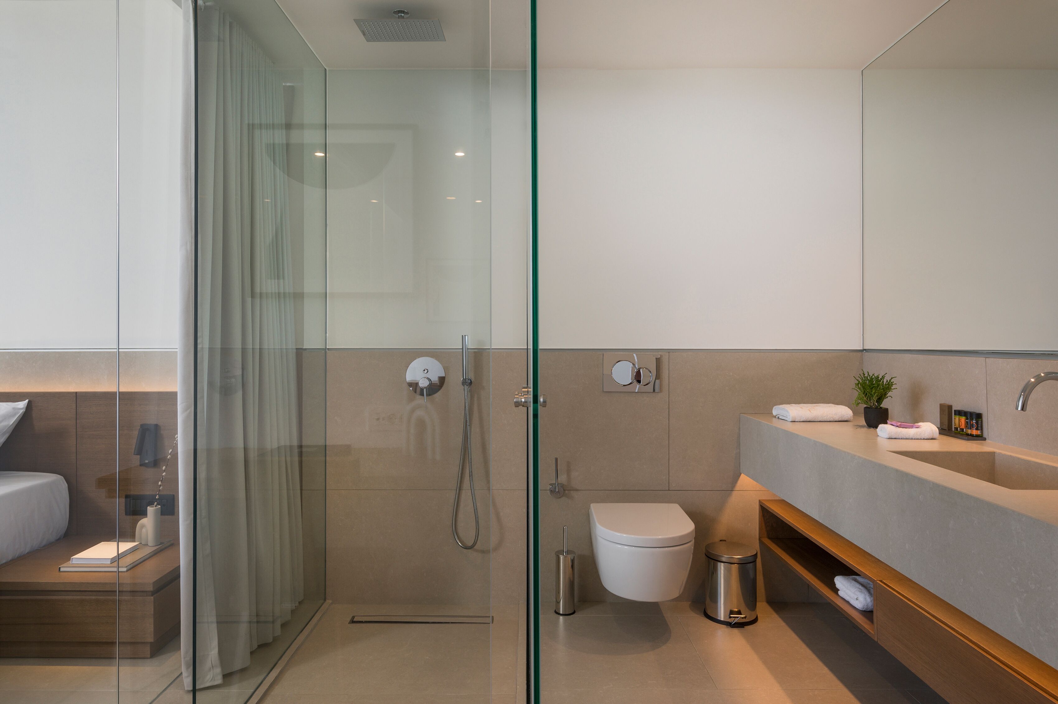 A modern bathroom with a glass shower enclosure, a toilet, a sink with a vanity, and a bedside table with a lamp.