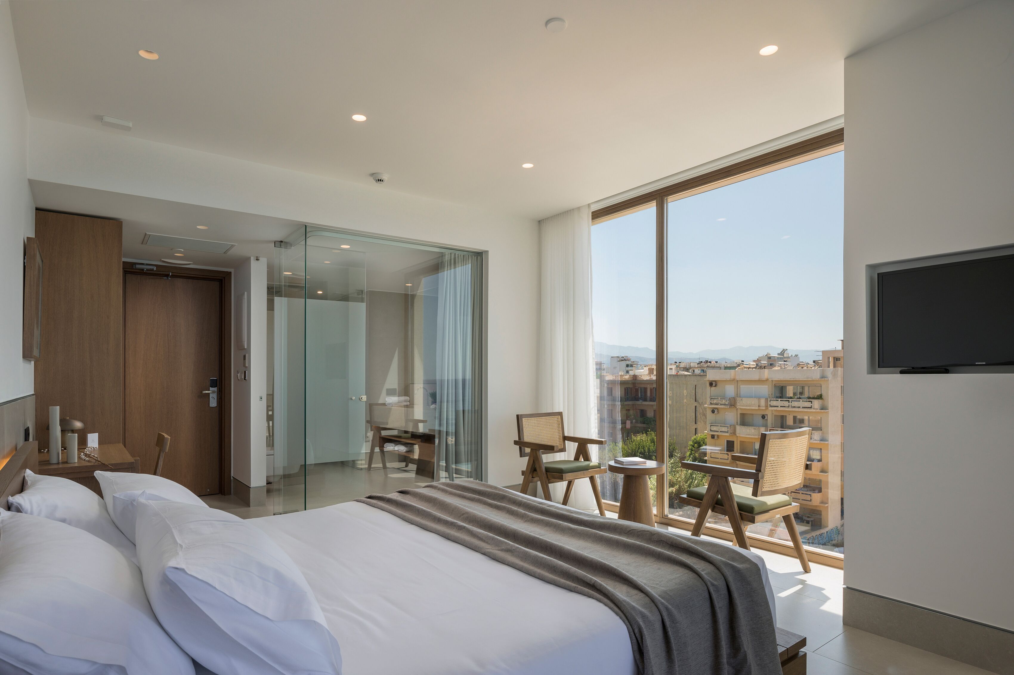 Une chambre d’hôtel moderne, dotée d’un grand lit, d’un bureau et d’une chaise. Un téléviseur est fixé au mur et la chambre comprend un balcon avec vue sur la ville.