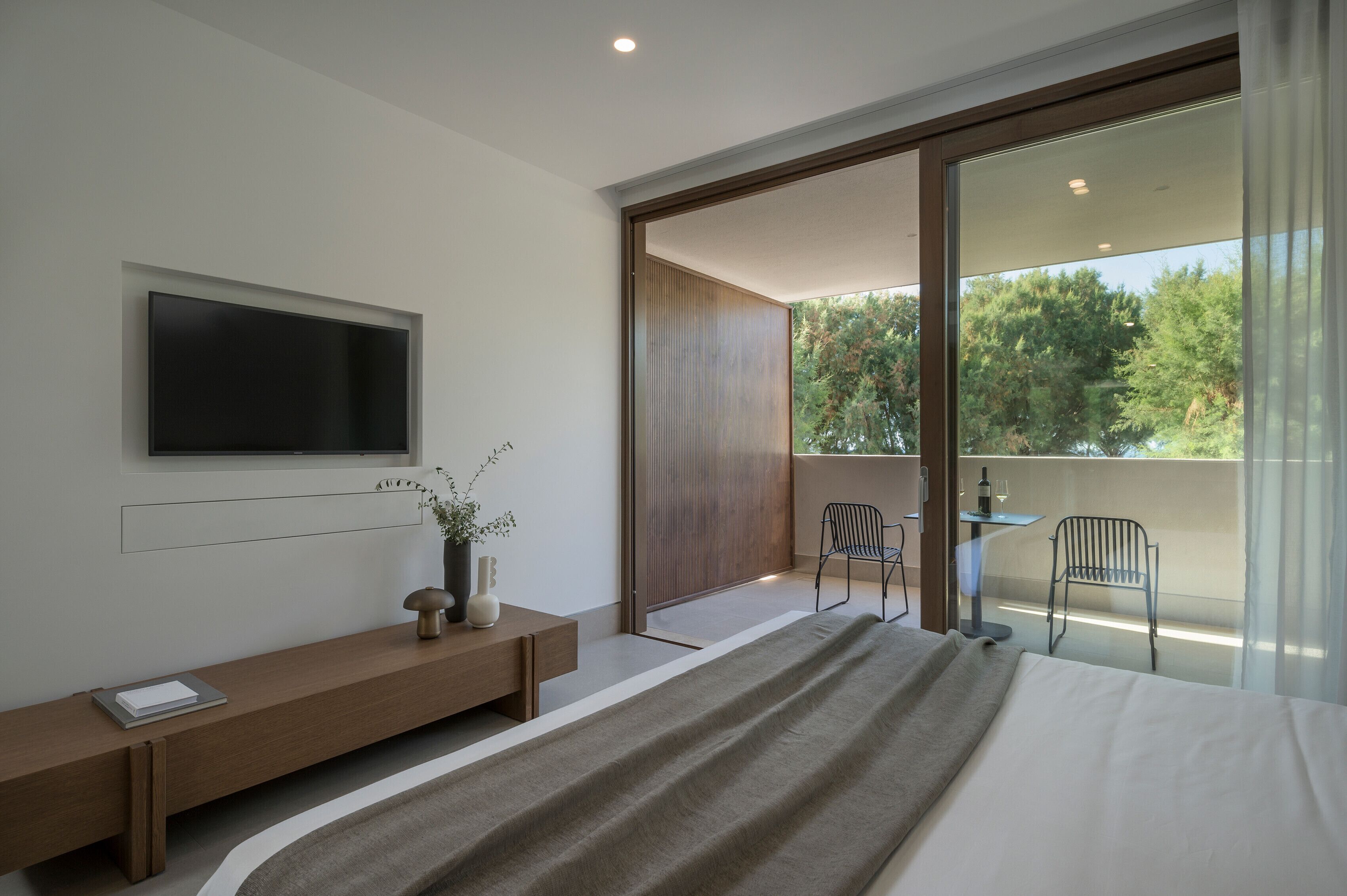 A modern bedroom with a large bed, a TV mounted on the wall, a bedside table with a vase, and a balcony with chairs and a table.