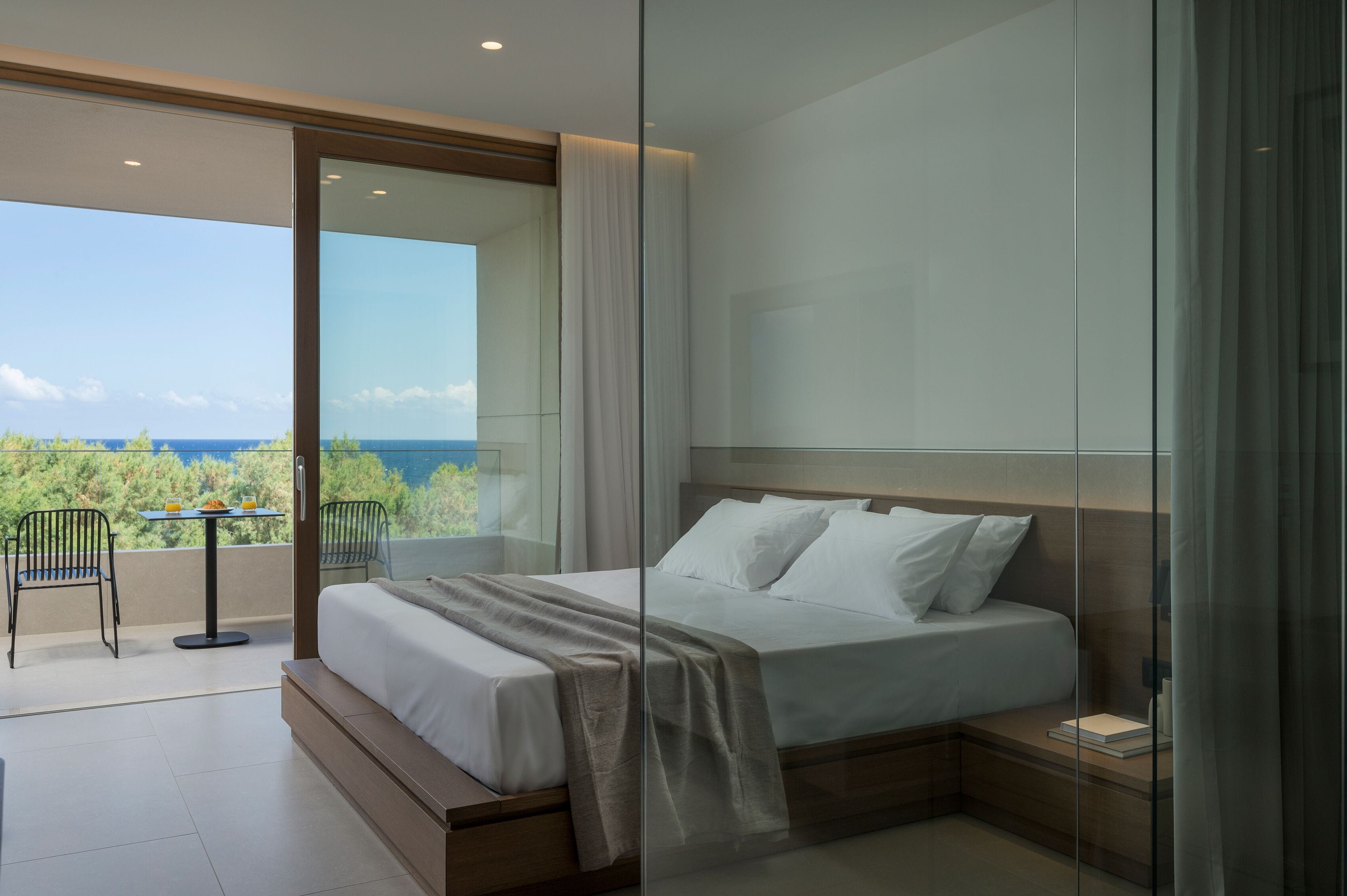 A modern bedroom with a large bed, a glass door leading to a balcony, and a view of the sea.