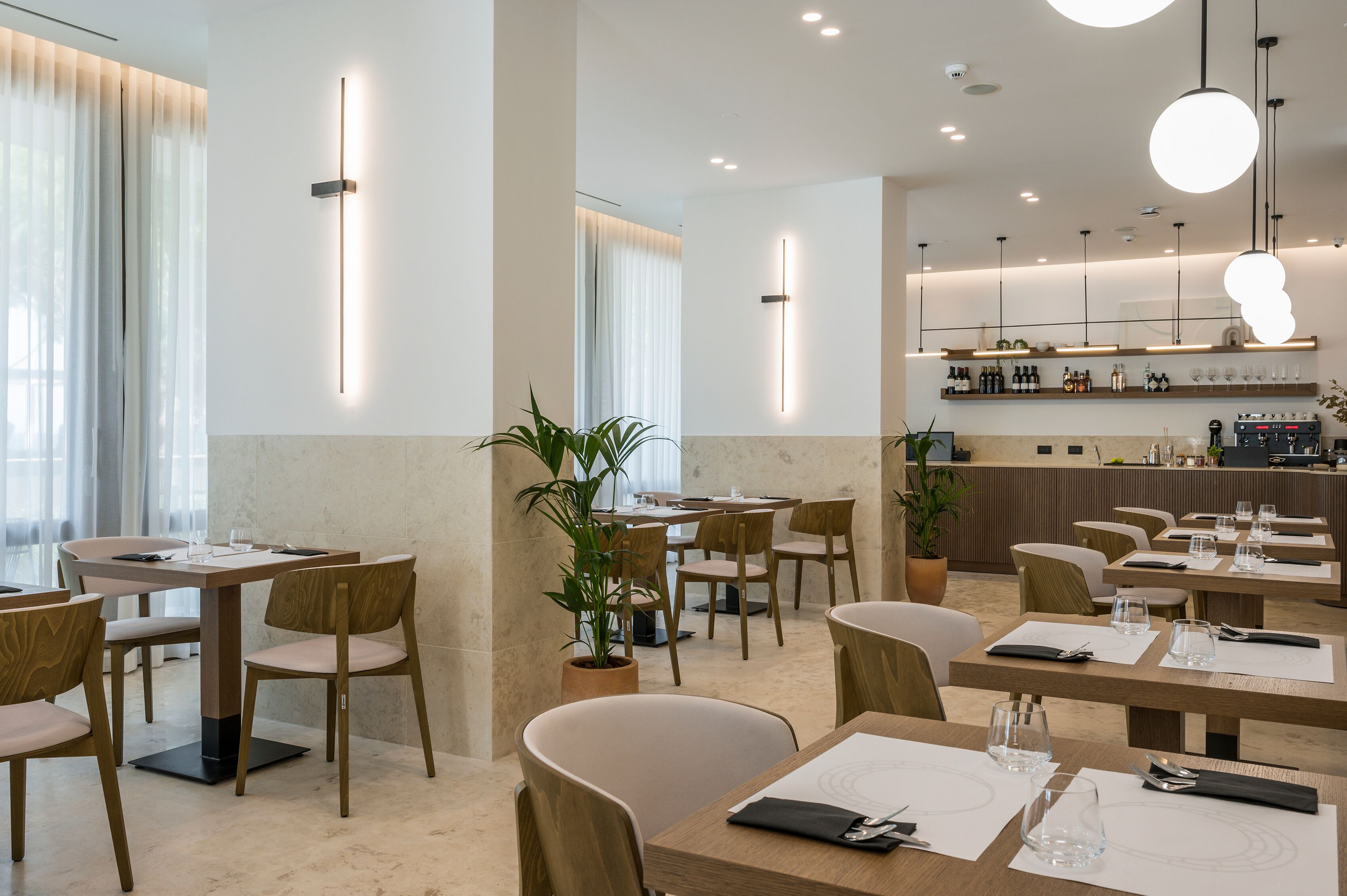 Un restaurant moderne avec des tables et des chaises en bois, un bar avec des étagères remplies de bouteilles et des luminaires suspendus.