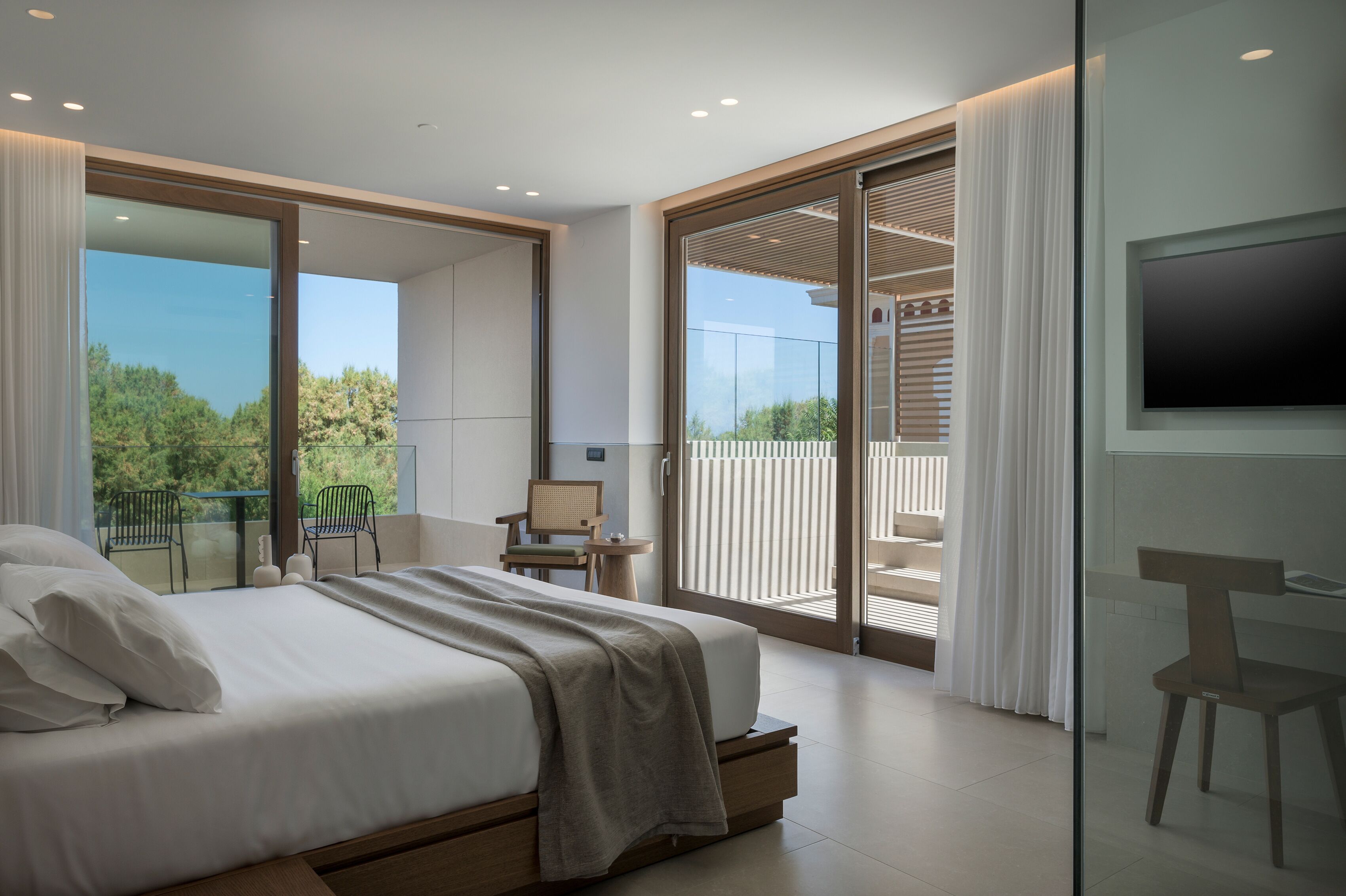 A modern bedroom with a large bed, a TV, and a balcony with a view of greenery.
