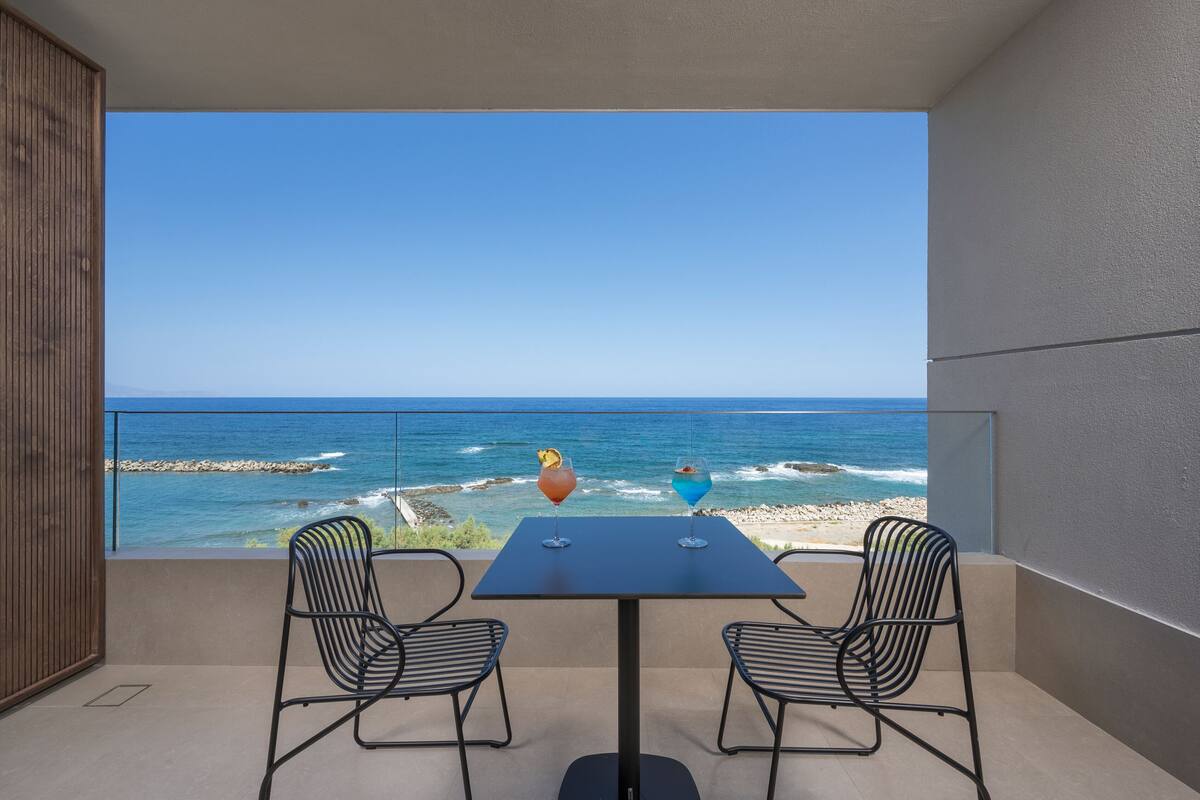 Ein Balkon mit Tisch und zwei Stühlen, mit Blick auf einen Strand, das Meer und eine felsige Küste.