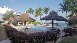 A pool area with lounge chairs and thatched umbrellas, a clear view of the ocean, and a building in the background.
