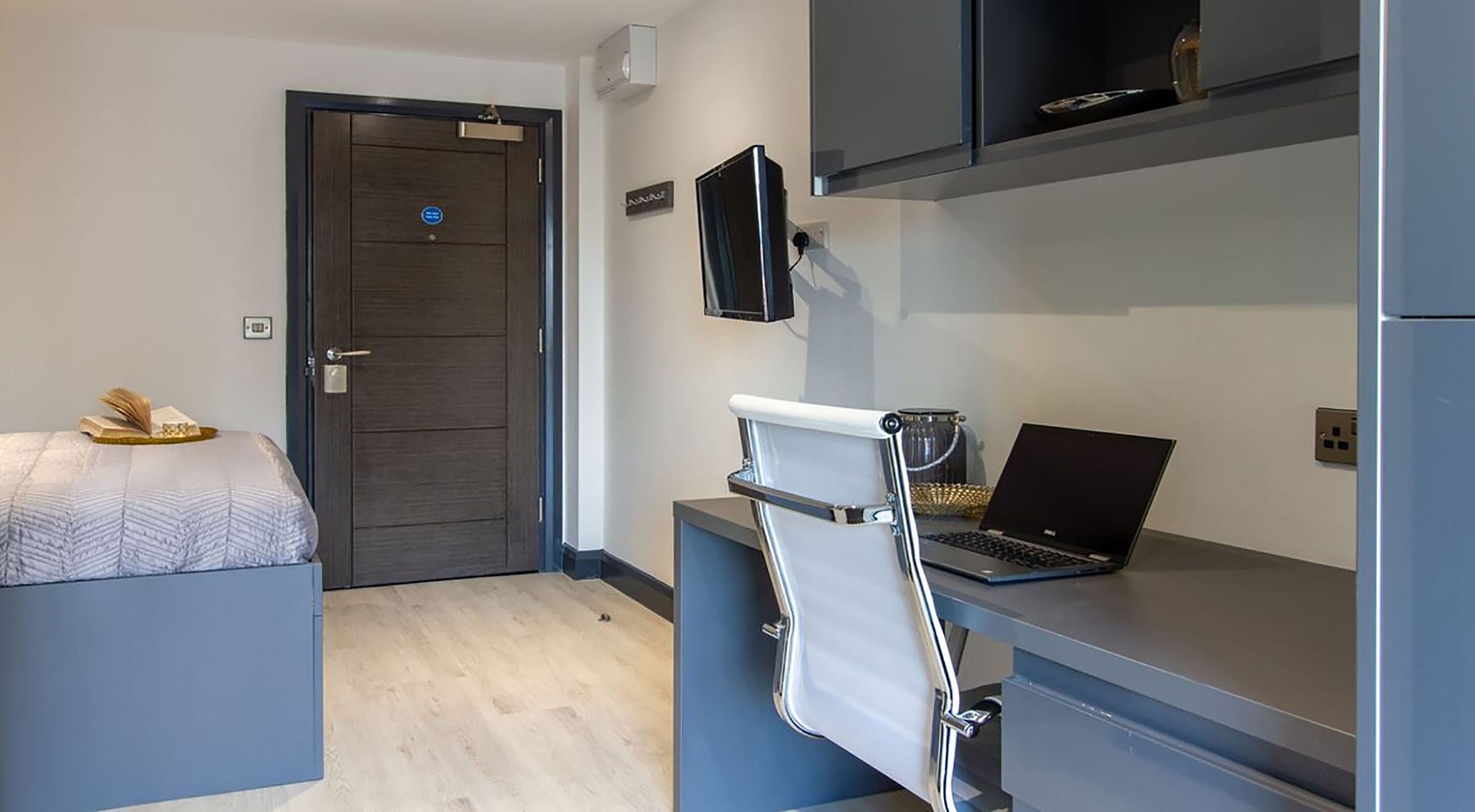 Ein kleines Hotelzimmer mit Bett, Schreibtisch mit Laptop, Stuhl und Wandfernseher.