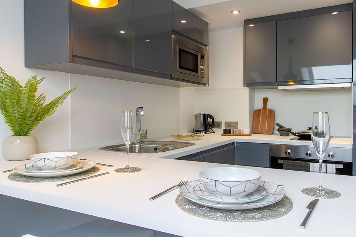 A modern kitchen with dark cabinets, a microwave, and a sink. There is a plant on the left and a dining area with plates and cutlery on the counter.