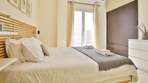 A neatly made bed with pillows and a blanket, a bedside table with a lamp, and a window with curtains. - Nautilus Apartments Airport by Airstay (Spata-Artemida)