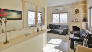 A living room with a sofa, a coffee table, and a fireplace. There are windows with crossbars, a mirror, and a vase with flowers. - Nautilus Apartments Airport by Airstay (Spata-Artemida)