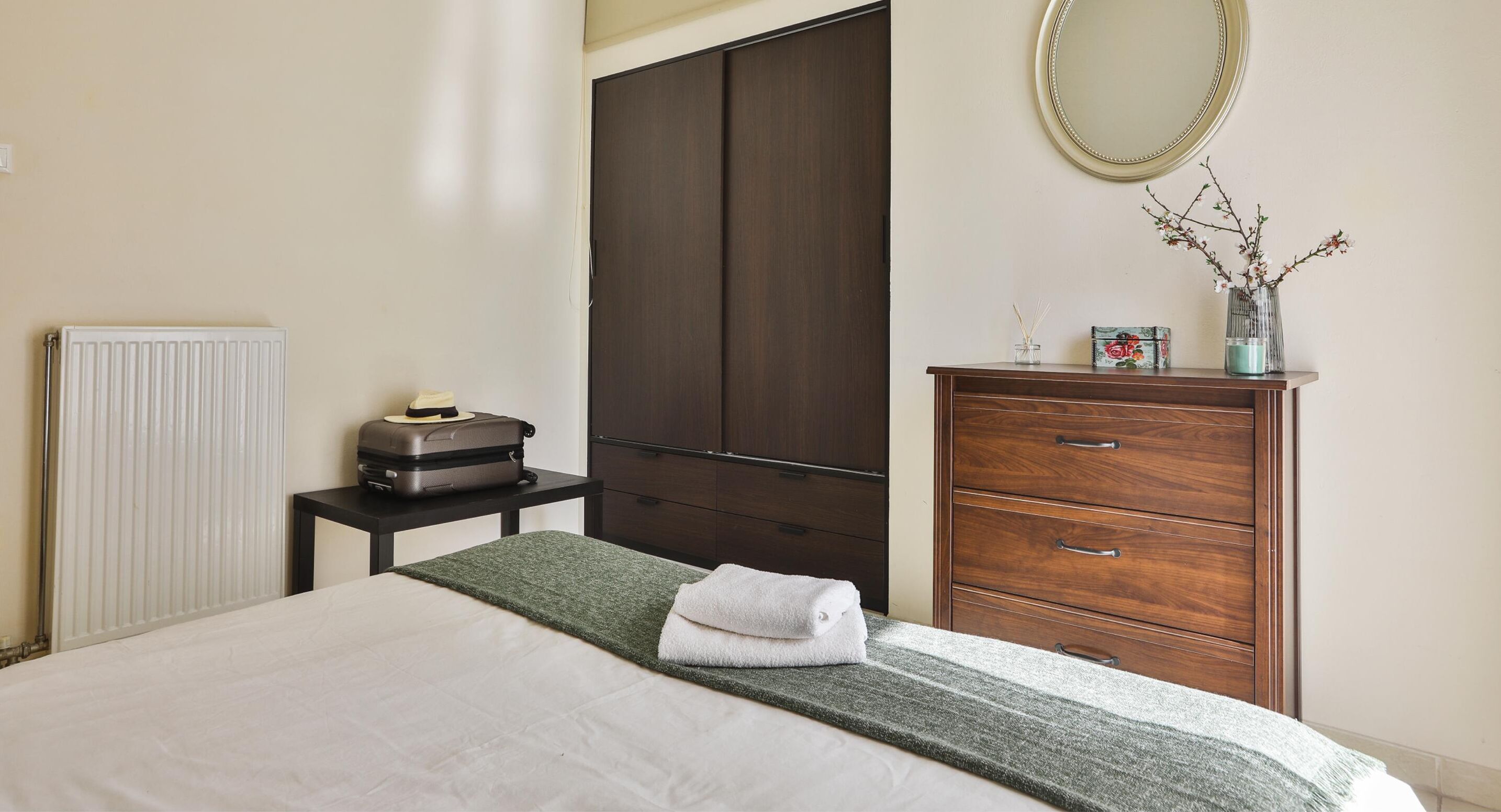 A hotel room with a bed, a dresser, a radiator, a suitcase, and a mirror on the wall.