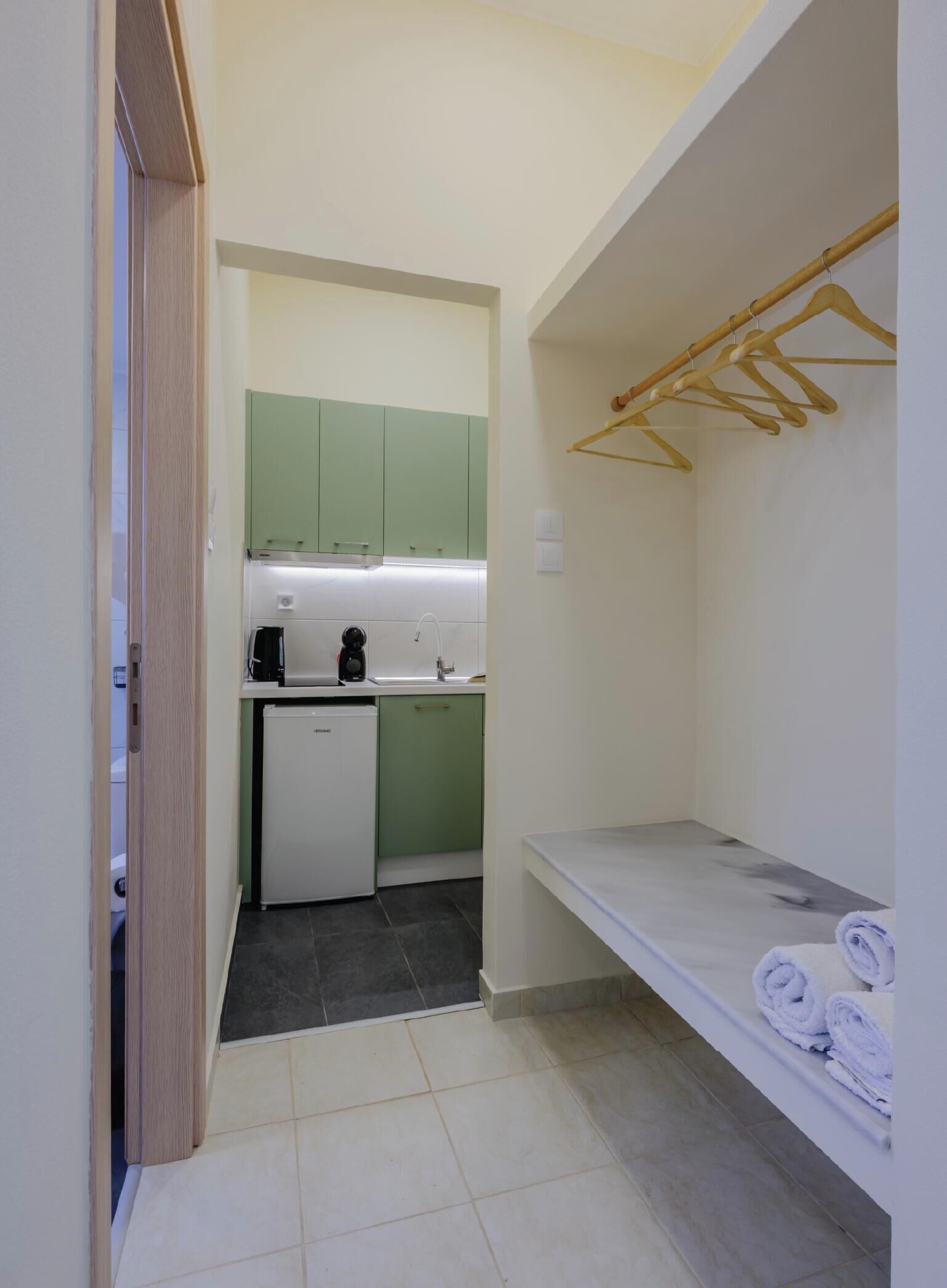 Een compacte keuken met een koelkast, een koffiezetapparaat en een spoelbak. Er is een klein plankje met handdoeken en een houten droogrek.