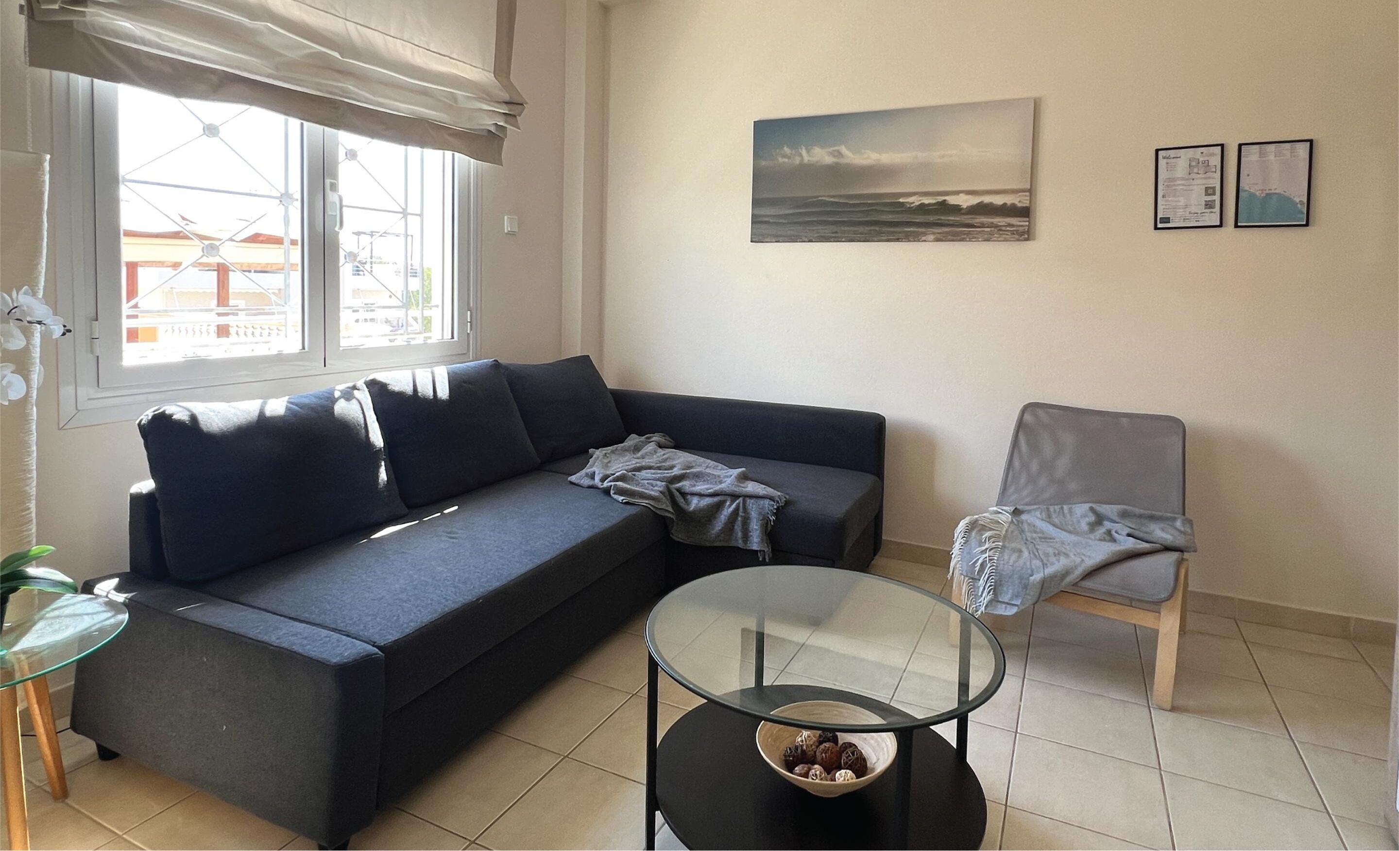 A living room with a sofa, a glass coffee table, and a chair. There is a window with a view of buildings outside.