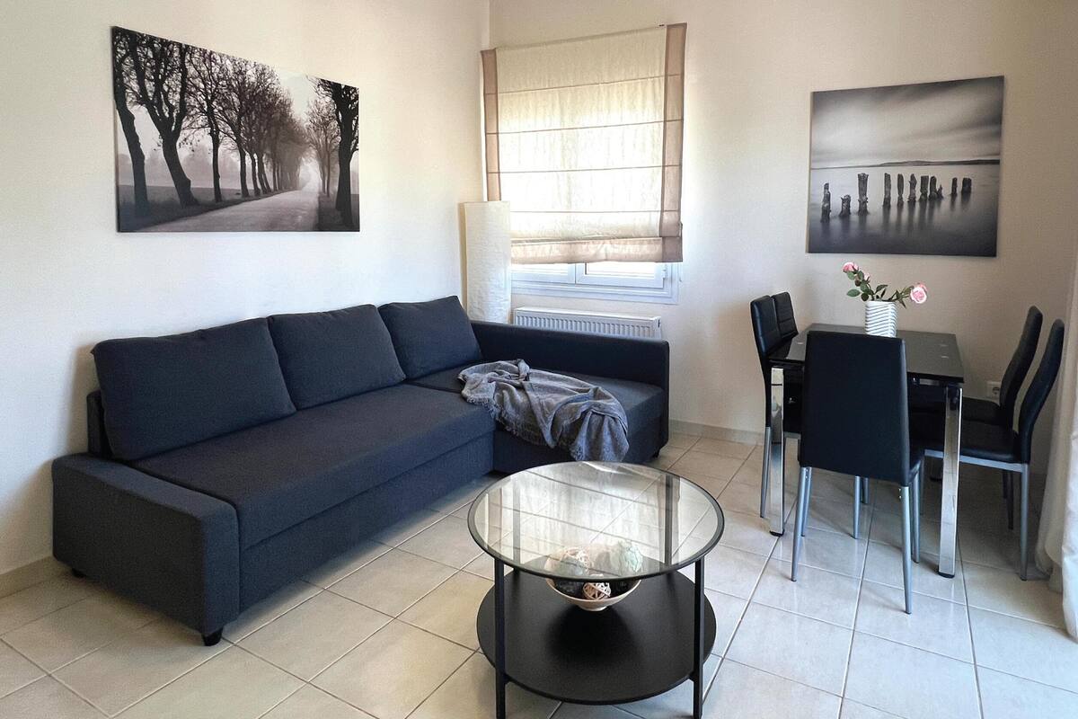 A modern living room with a dark sofa, a glass coffee table, a dining table with chairs, and two wall-mounted artworks.