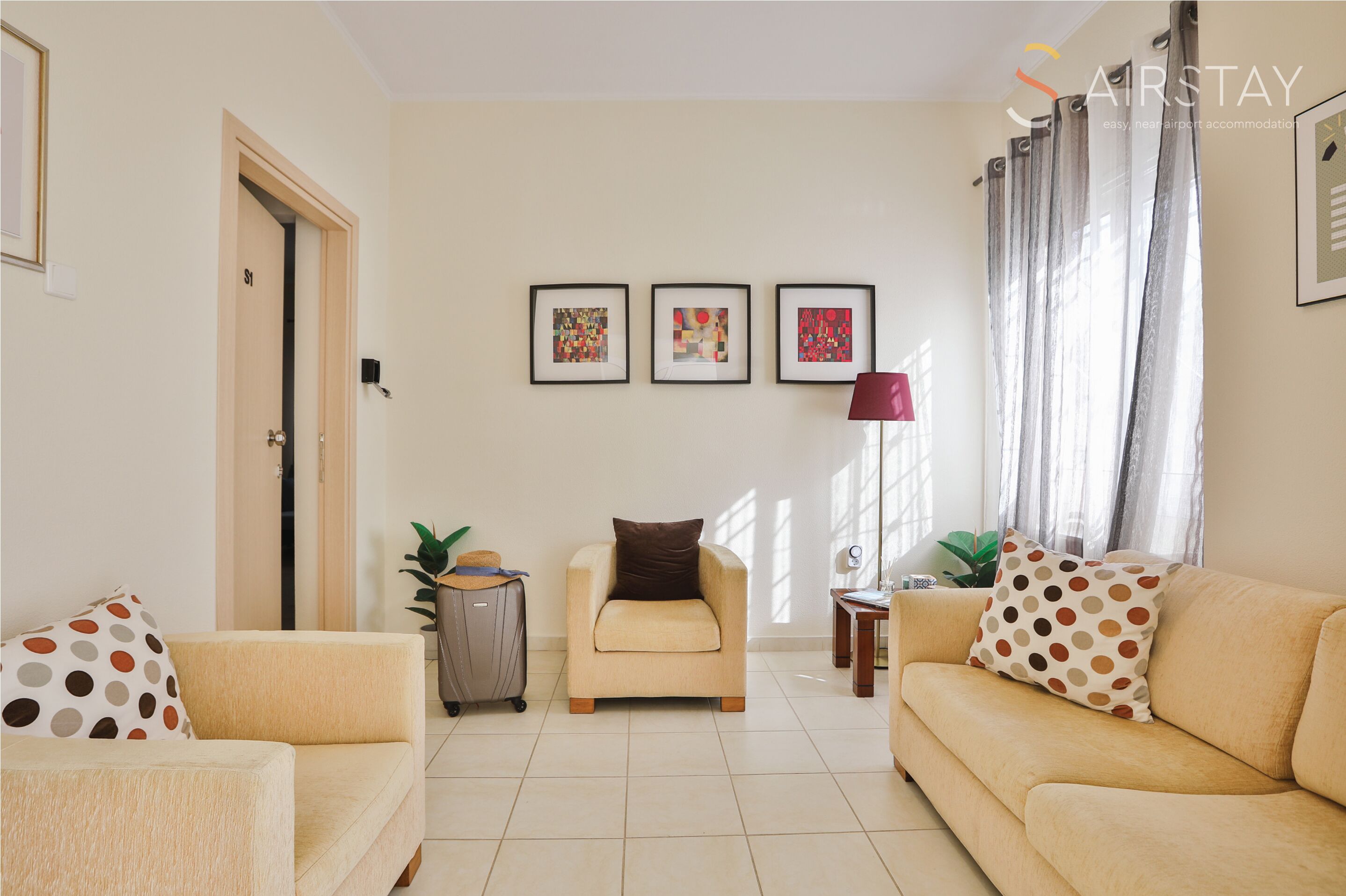 A living room with a beige sofa, a small armchair, and a coffee table. There are three framed pictures on the wall, a door, and a window with curtains.
