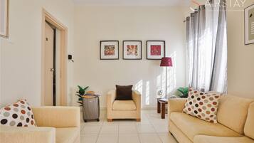 A living room with a beige sofa, a small armchair, and a coffee table. There are three framed pictures on the wall, a door, and a window with curtains.