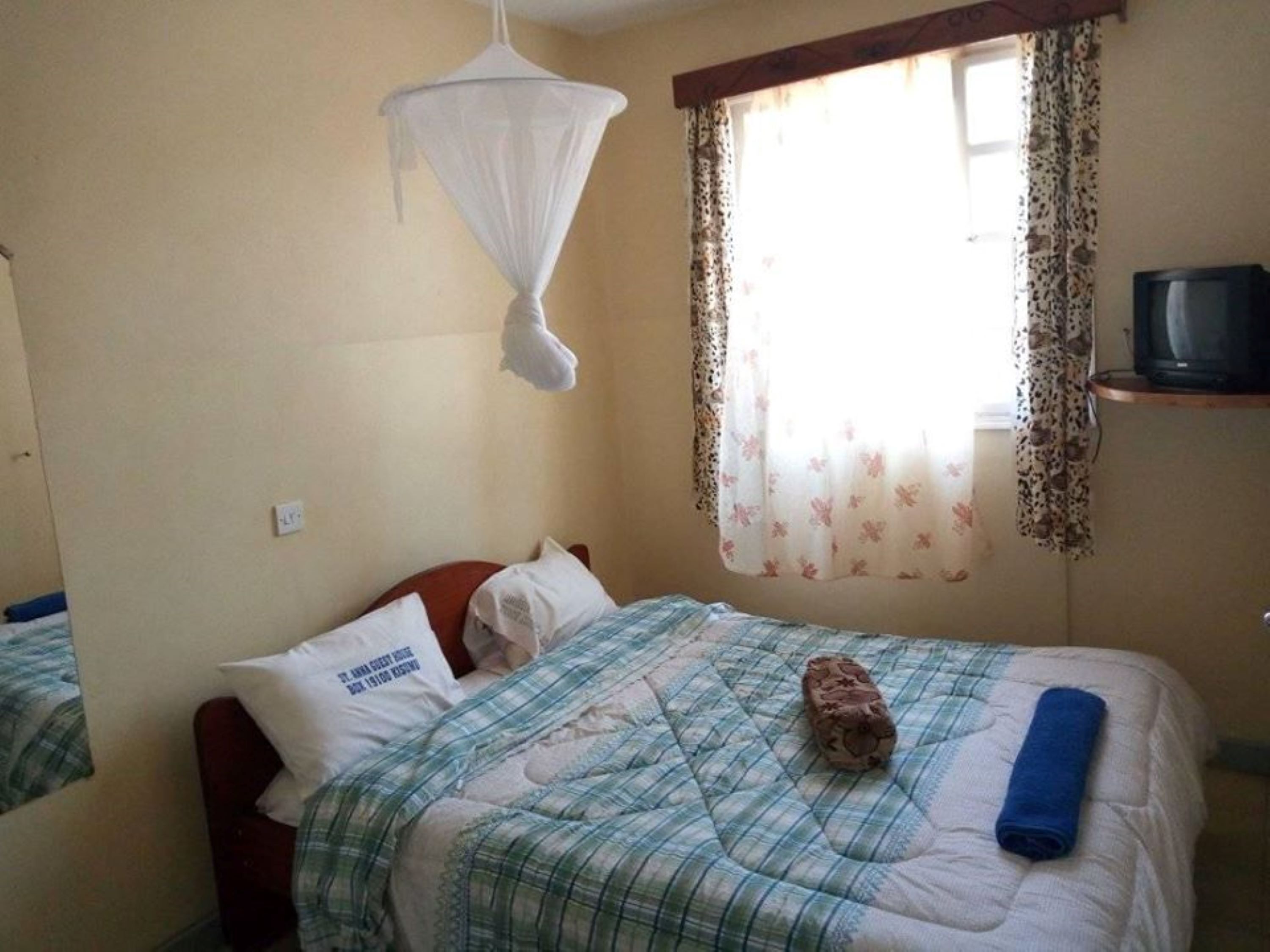 Un lit simple avec une couverture à carreaux, une serviette enroulée et un oreiller orné d’un motif. Une moustiquaire est suspendue au-dessus du lit.