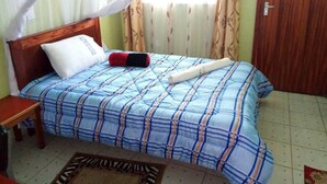 A bedroom with a bed, a wooden headboard, a bedside table, a rolled-up towel, and a window with curtains. - St. Anna Guest House (Kisumu)