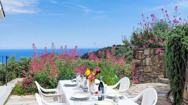 Une table dressée pour le dîner, avec vue sur la mer, entourée de fleurs roses et de verdure.