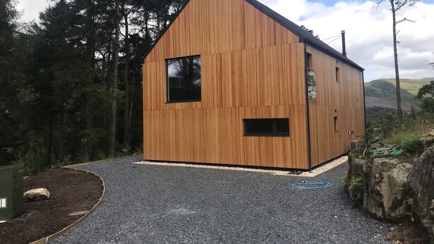 A wooden cabin with a sloped roof, a gravel driveway, and a forested backdrop. - An Caisteal Lodge With Cascading Waterfall View (Crianlarich)