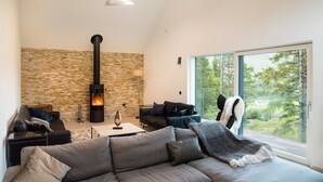 A modern living room with a stone wall, a fireplace, and a large window offering a view of trees. - An Caisteal Lodge With Cascading Waterfall View (Crianlarich)