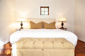 A neatly made bed with a headboard, two bedside lamps, a small window, and a bench at the foot of the bed.