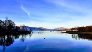 A calm lake with clear reflections, surrounded by trees and mountains under a blue sky. - The Wee Studio For Two (Stirling)