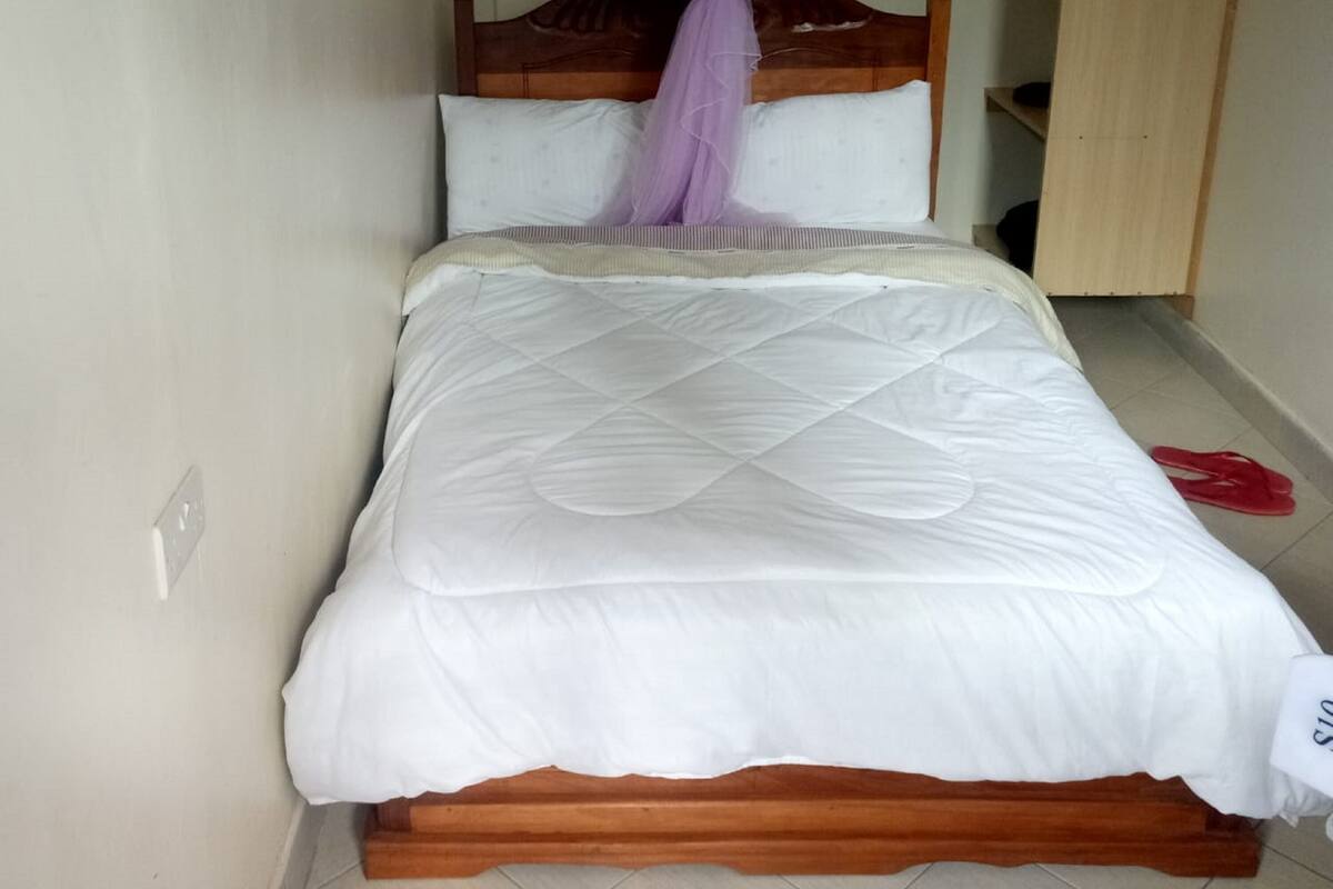 Uma cama de solteiro com estrutura de madeira e colchão branco acolchoado. Um dossel roxo está amarrado na cabeceira da cama.