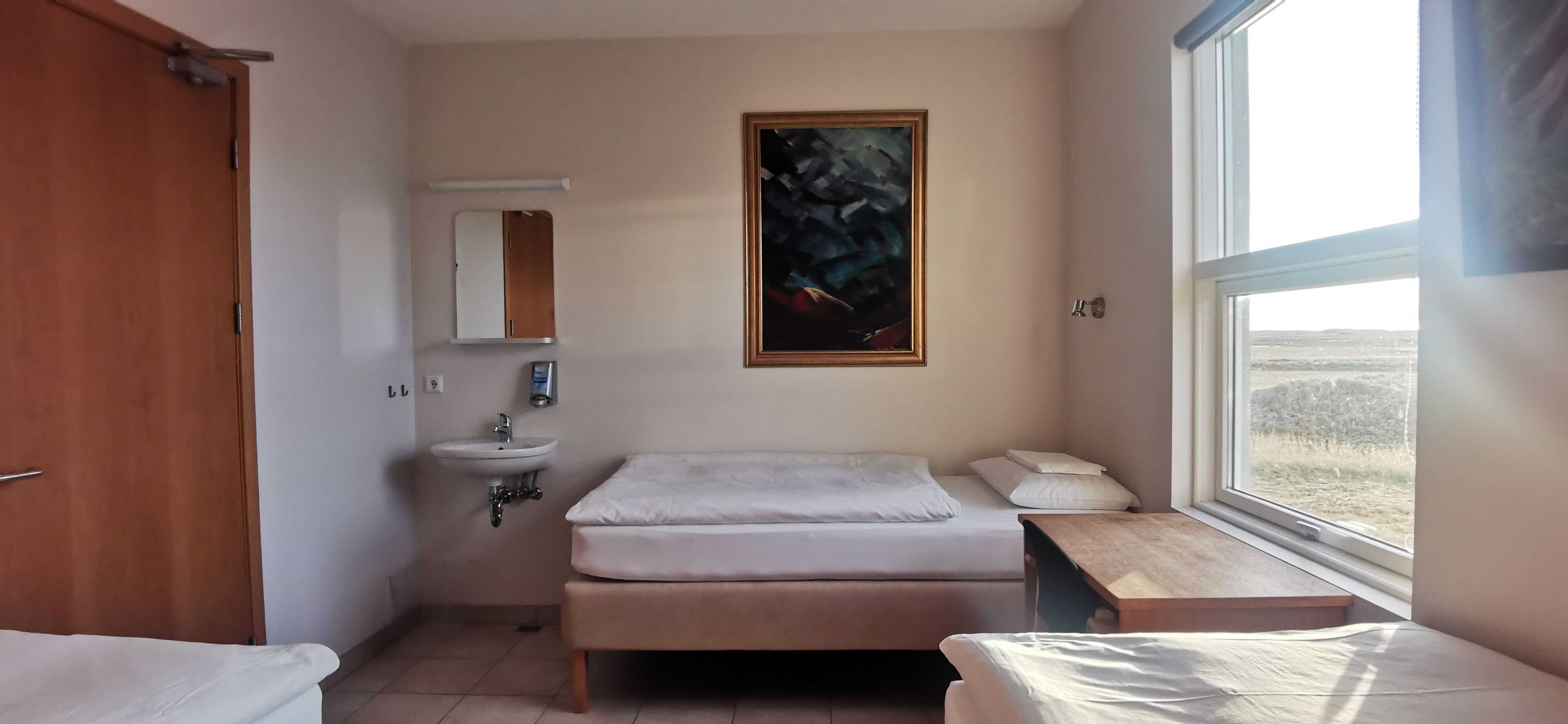 A small, clean room with a bed, a bedside table, a window with a view of the outdoors, a sink, and a mirror.