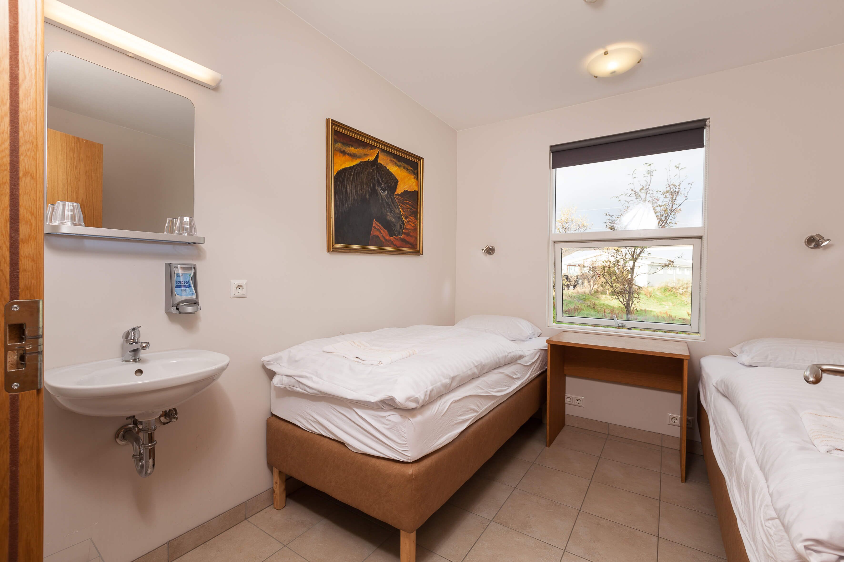 A bedroom with a bed, a bedside table, a window, and a bathroom with a sink and mirror.