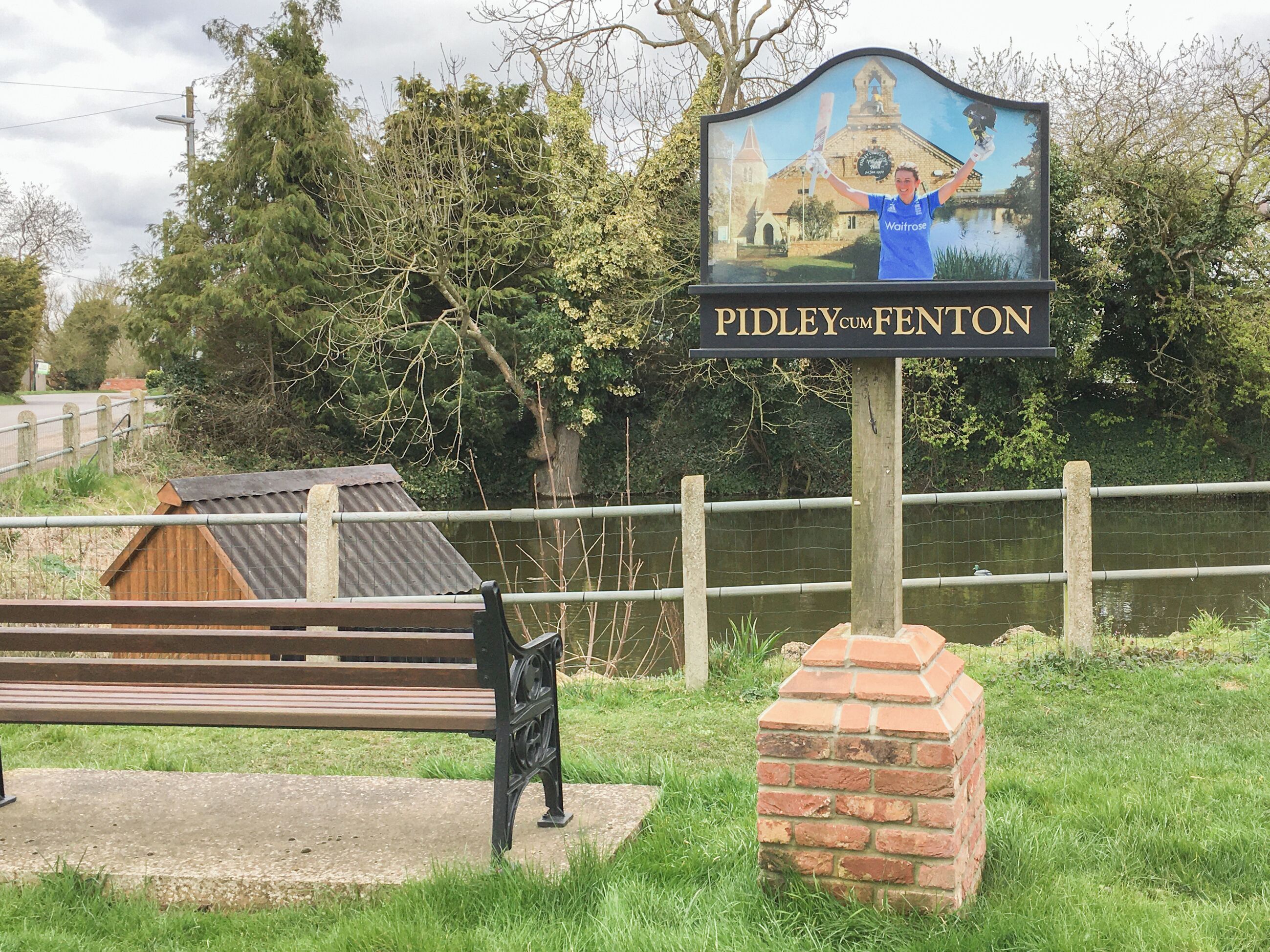 Schild für 'Pidley cum Feniton' mit Darstellung eines Gebäudes und einer Person, die einen Cricket-Schläger hält.