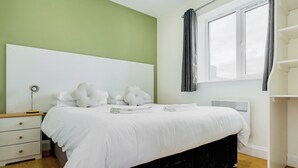 Una cama bien hecha con ropa blanca, una lámpara de noche y una ventana con cortinas.