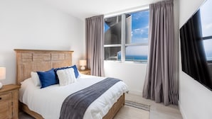 A bedroom with a large bed, a wooden headboard, a nightstand, a lamp, a window with curtains, and a view of the sea. - 43rd Floor Penthouse Ocean Views Private Terraces (Hollywood)
