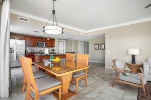 A modern kitchen with stainless steel appliances, a dining area with a glass table and wooden chairs, and a living area with a sofa and lamp.