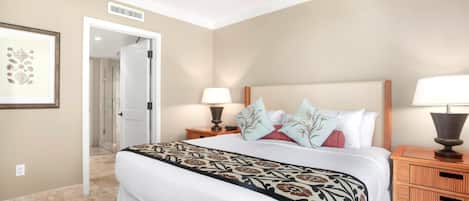 Une chambre d’hôtel avec un grand lit, deux tables de chevet avec des lampes, un ventilateur de plafond et une porte donnant sur une autre pièce.