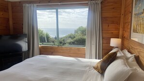 A bedroom with a large bed, a TV, and a window with curtains. - Seaview, Sun and Surf - Piha Holiday Home (Piha)