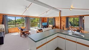 A modern cabin with a kitchen, dining area, and a balcony with a view of the forest. - Seaview, Sun and Surf - Piha Holiday Home (Piha)